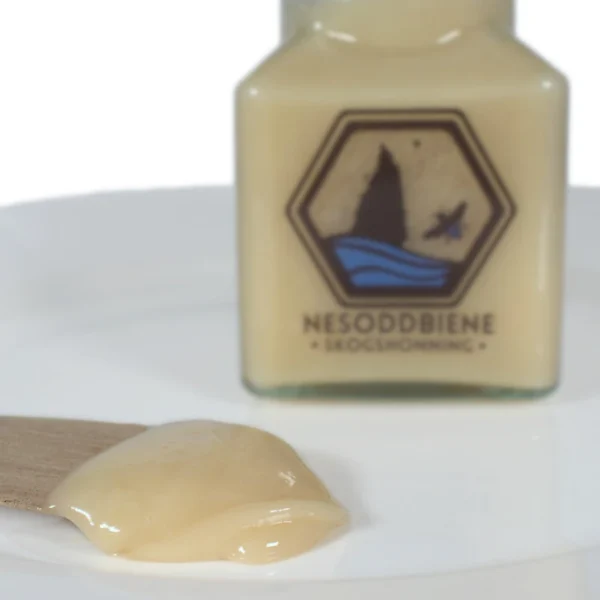Image showing a wooden spreader knife with our Forest Honey on it. The honey is creamed, or soft set. In the background you see a jar of the same honey.